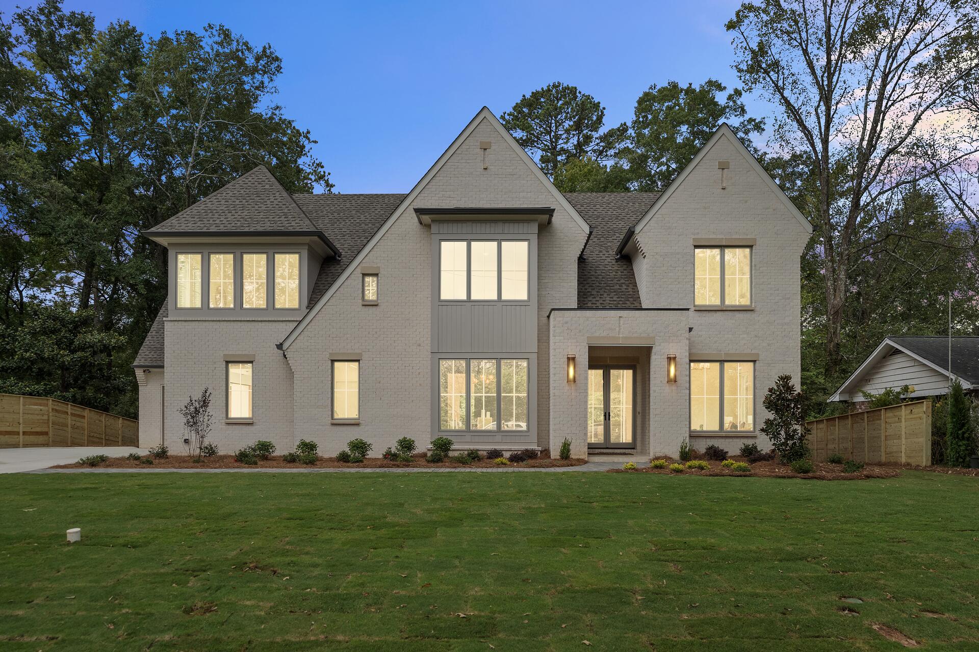 A shingle-clad custom home with a white wraparound porch, set back on a mown lawn in autumn light